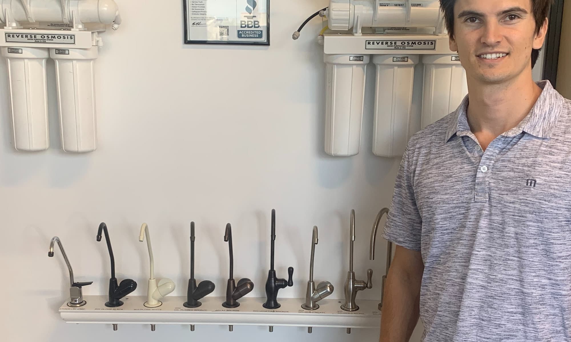 An AquaCo team member beside two reverse osmosis systems and a display of drinking water faucet finishes